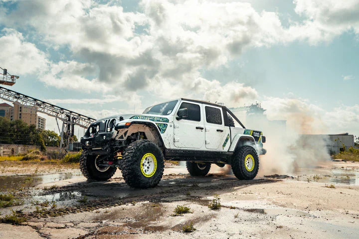 Lifted Jeep Gladiator with off-road modifications, including oversized tires, fender flares, and custom hood decals, kicking up dust in an industrial setting.